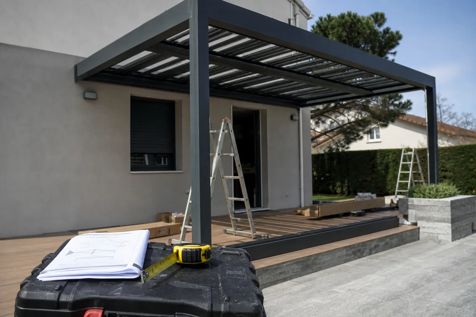 Pergola bioclimatique en cours d’installation, mètre ruban et notice de pose sur une caisse à outils