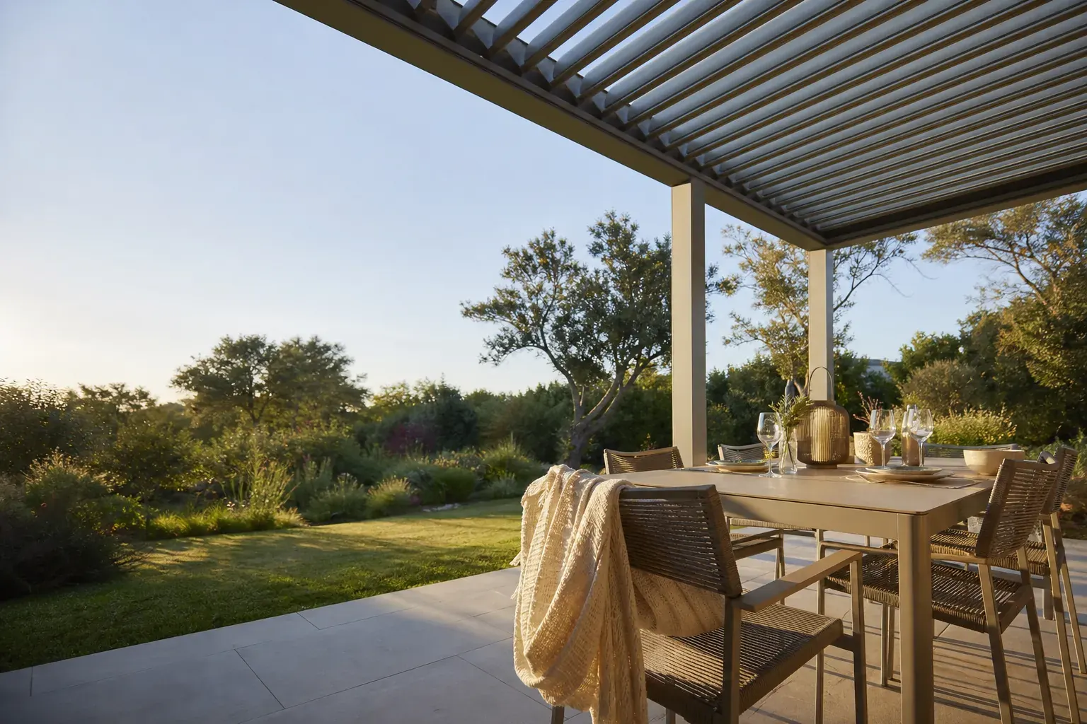 Terrasse habitée sous une pergola bioclimatique, lames mi-ouvertes, table dressée en fin de journée